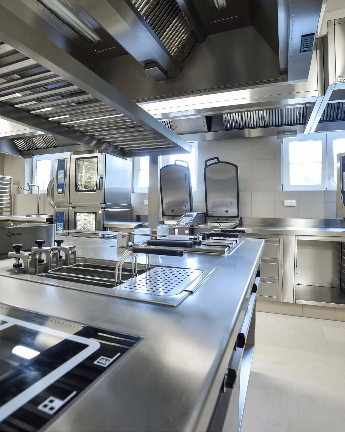 Modern commercial kitchen with stainless steel countertops, cooking equipment, and large ventilation hoods.